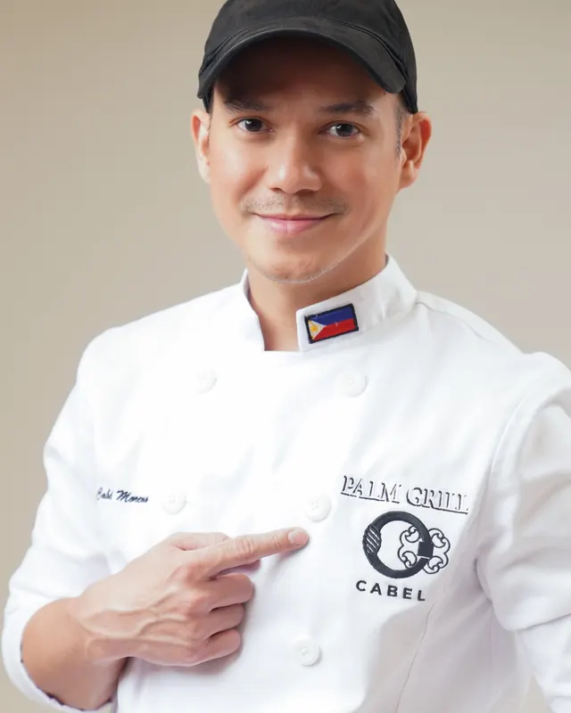 Chef Miggy Cabel Moreno in chef's whites, pointing to the Palm Grill and Cabel logos on his jacket